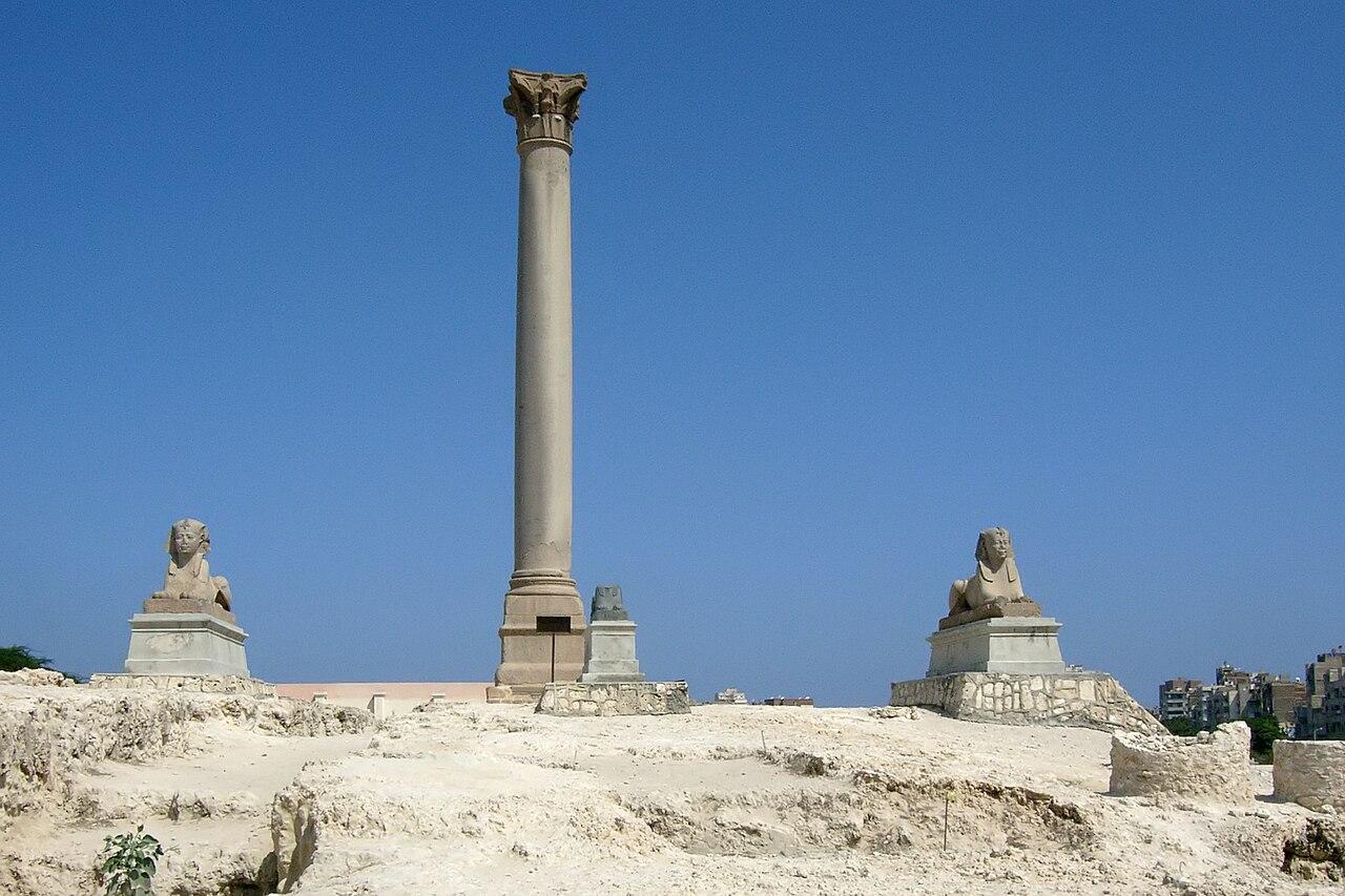 Pompey's Pillar rising above ancient ruins in Alexandria, Egypt, with modern apartment buildings and clear blue sky behind