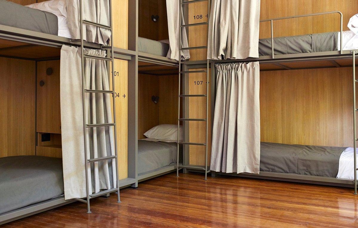 Rows of illuminated Japanese capsule hotel pods with white bedding, stacked two levels high in a dark modern interior