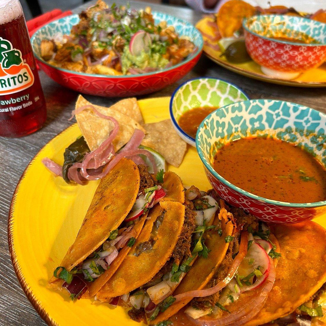 Hand holding a crispy corn tortilla taco filled with red adobo beef, guacamole, diced onion, and cilantro