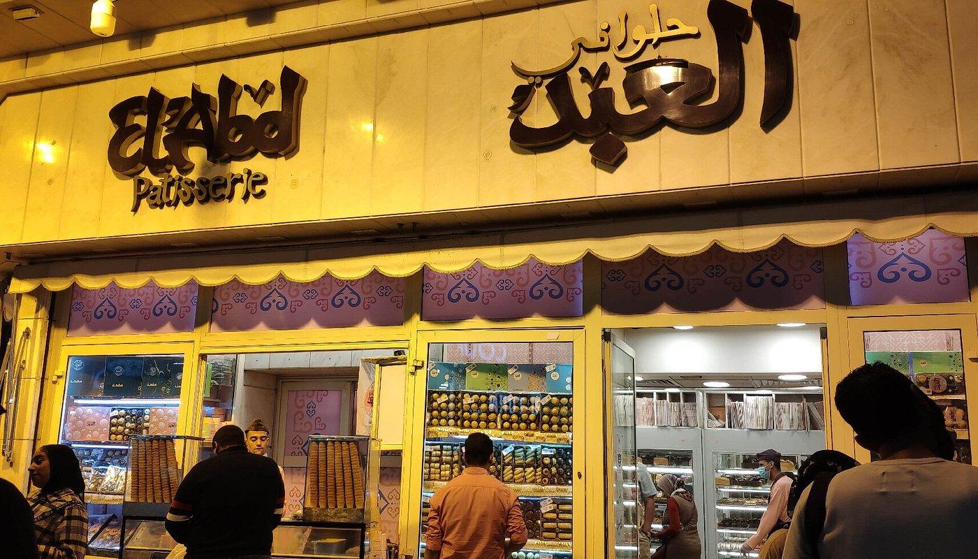 Busy Cairo street lined with parked cars and Arabic shop signs, leading to a stone church with a cross-topped spire