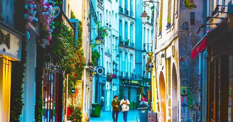 Narrow cobblestone alley in Le Marais Paris with tall Haussmann buildings, street lamps, and pedestrians passing bistro signs