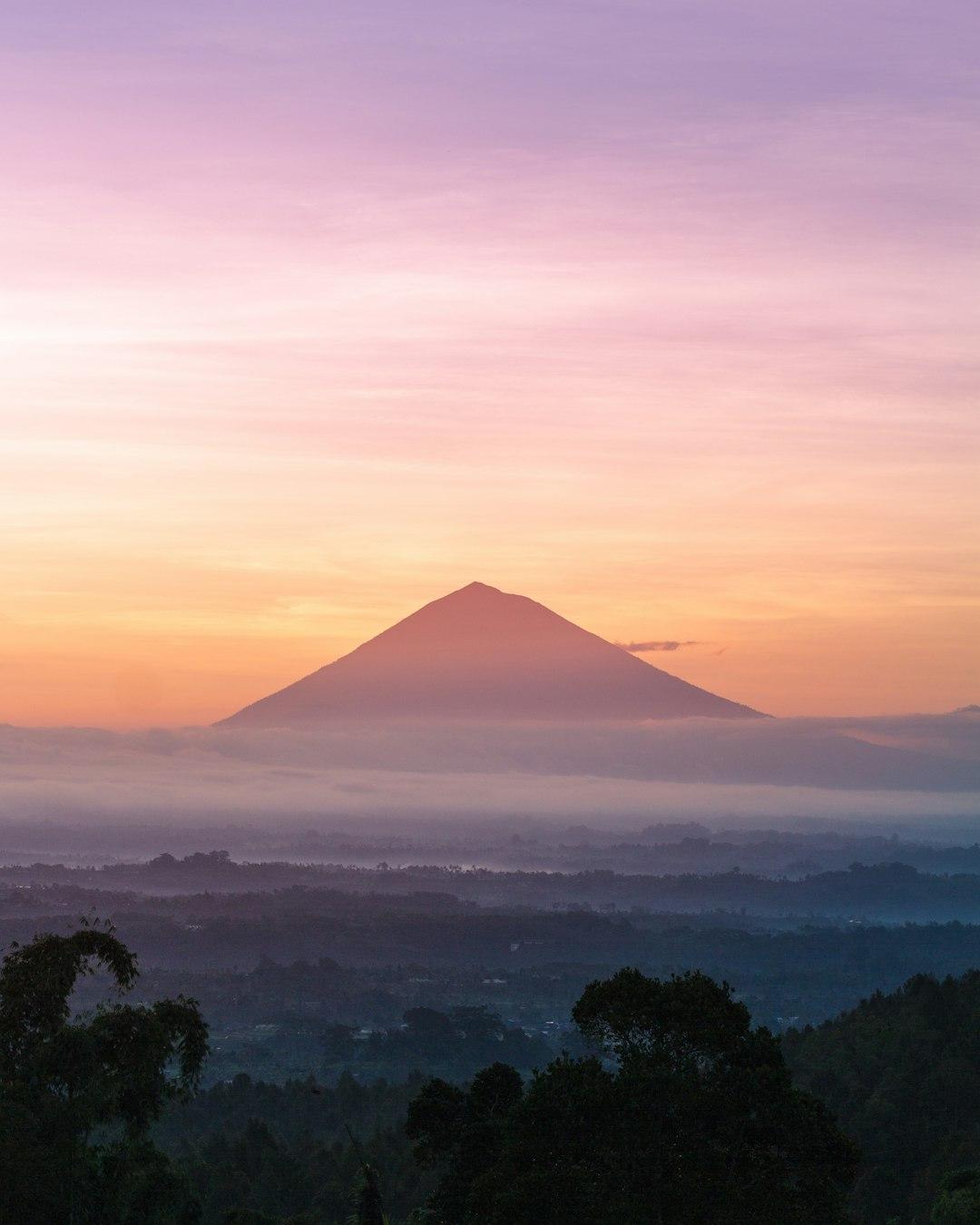 Silhouette of Mount Batur rising above misty valleys at sunrise, with pink, orange, and purple sky over Bali