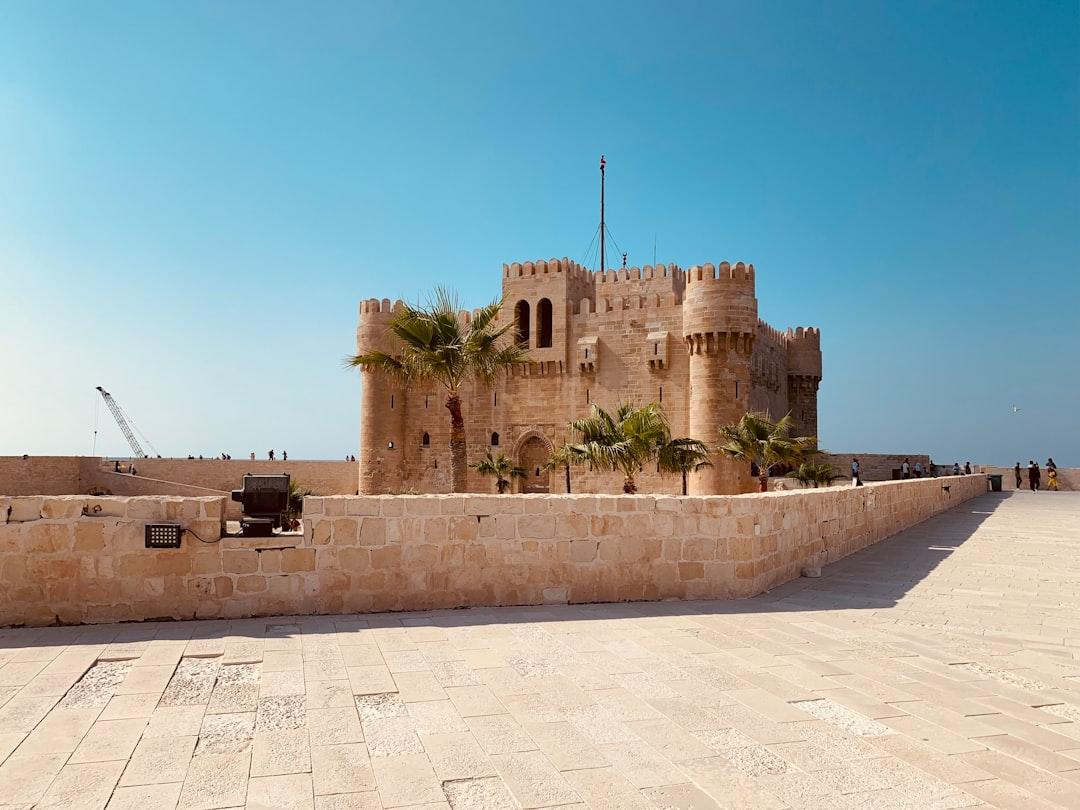 Qaitbay Citadel in Alexandria, Egypt — a sandy stone fortress with crenellated towers and palm trees under a clear blue sky