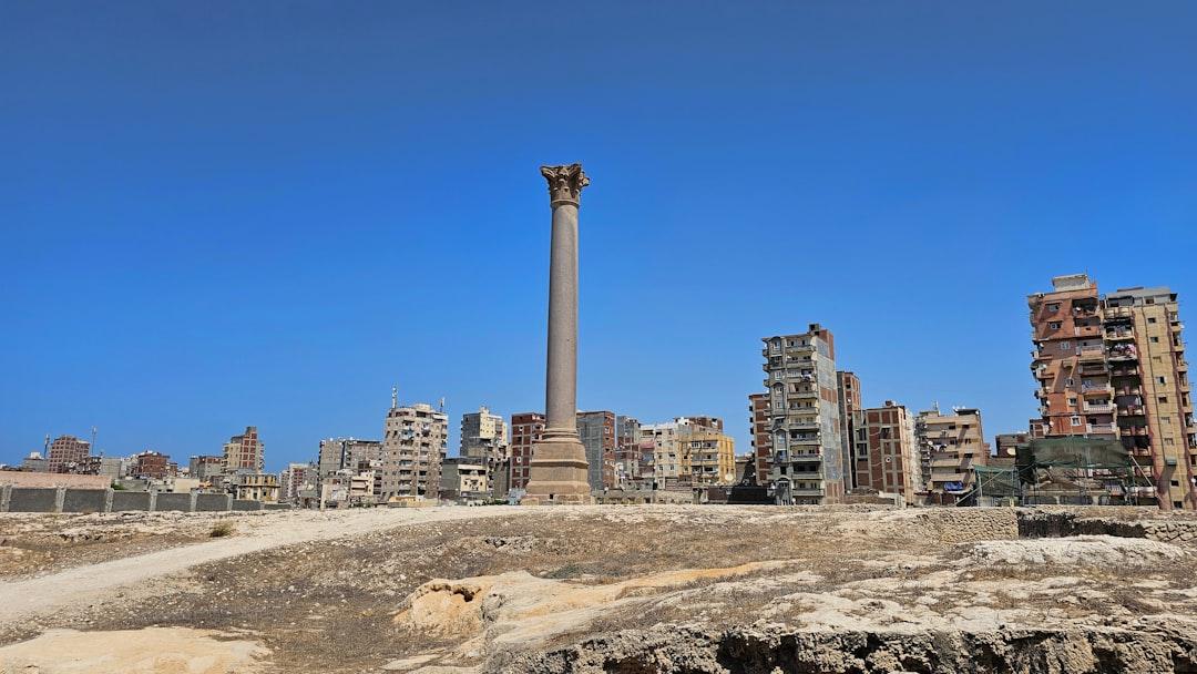 Pompey's Pillar rising above ancient ruins in Alexandria, Egypt, with modern apartment buildings and clear blue sky behind