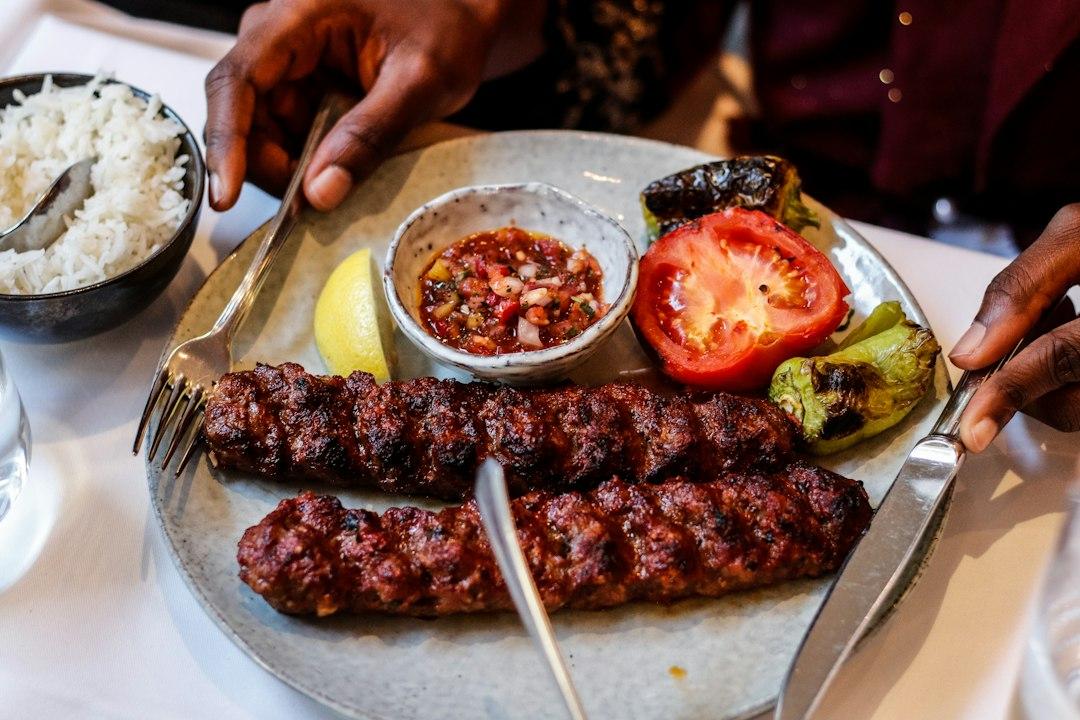 Grilled kofta kebabs on a plate with charred tomato, green pepper, salsa dip, lemon wedge, and a side bowl of white rice
