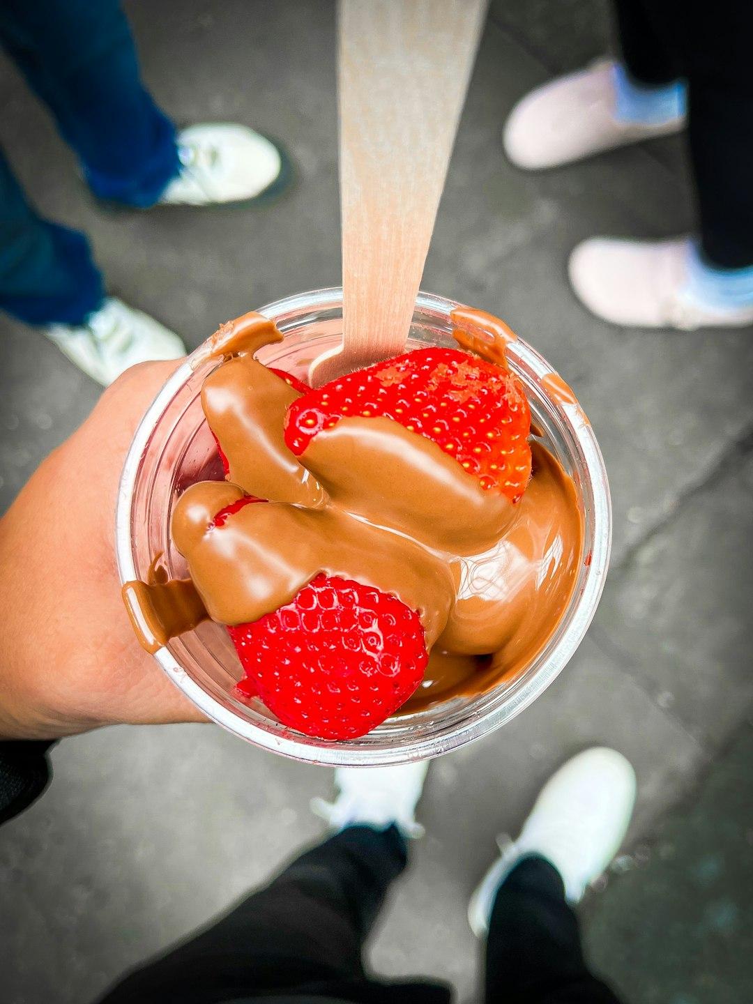 Hand holding a clear cup of fresh strawberries drizzled with melted chocolate and a wooden spoon at Borough Market