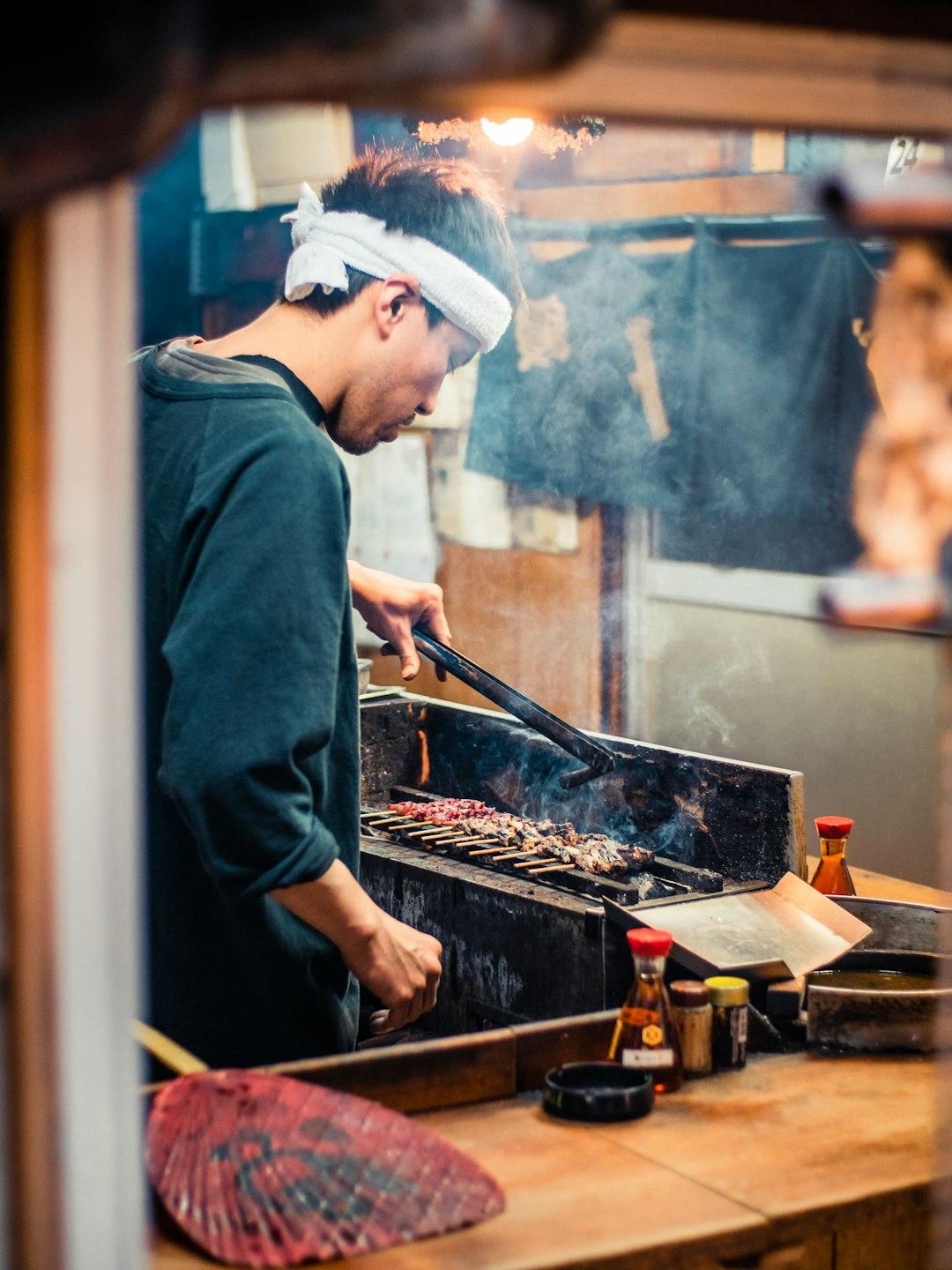 Japanese cook wearing a headband grills yakitori skewers over a smoky charcoal grill in a rustic izakaya kitchen