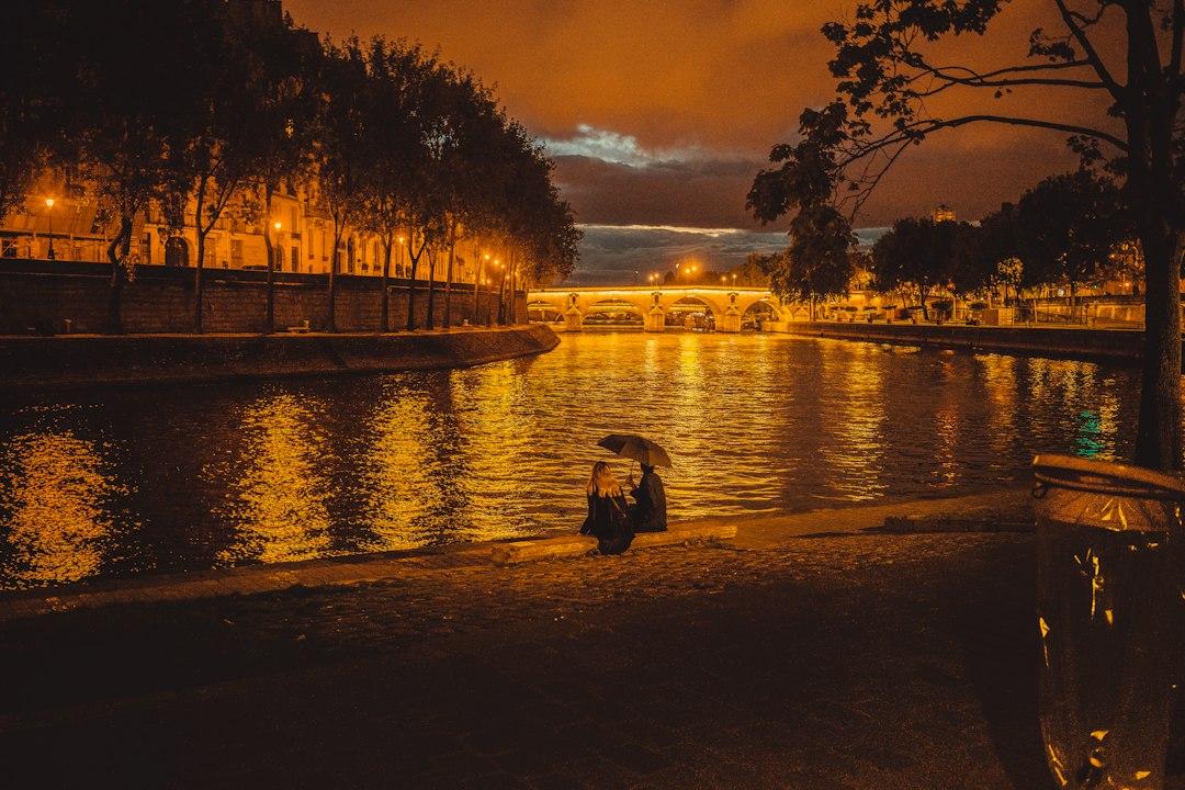Couple sharing an umbrella on the Seine riverbank at night, golden city lights reflecting on the water with an illuminated Paris bridge