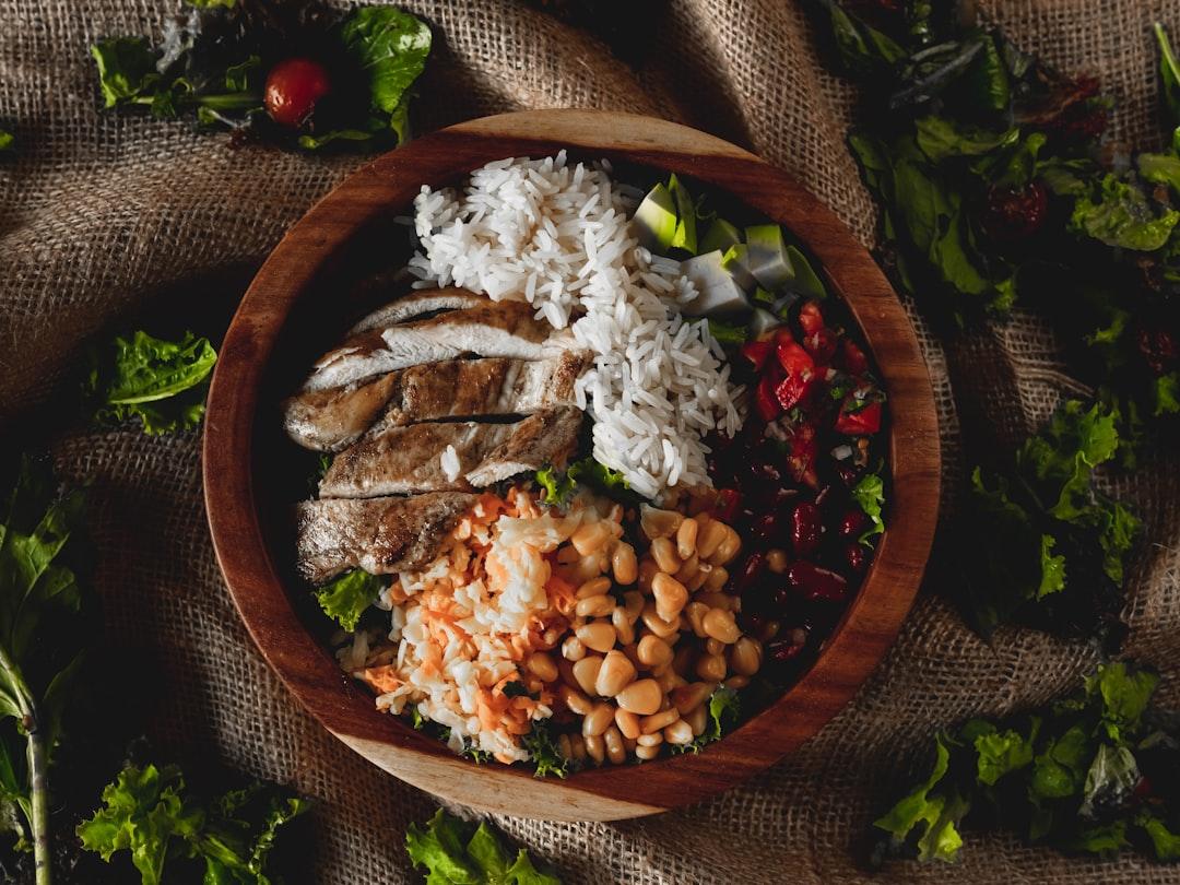 Wooden bowl filled with white rice, grilled chicken slices, white beans, red kidney beans, tomato salsa, and seasoned rice on burlap