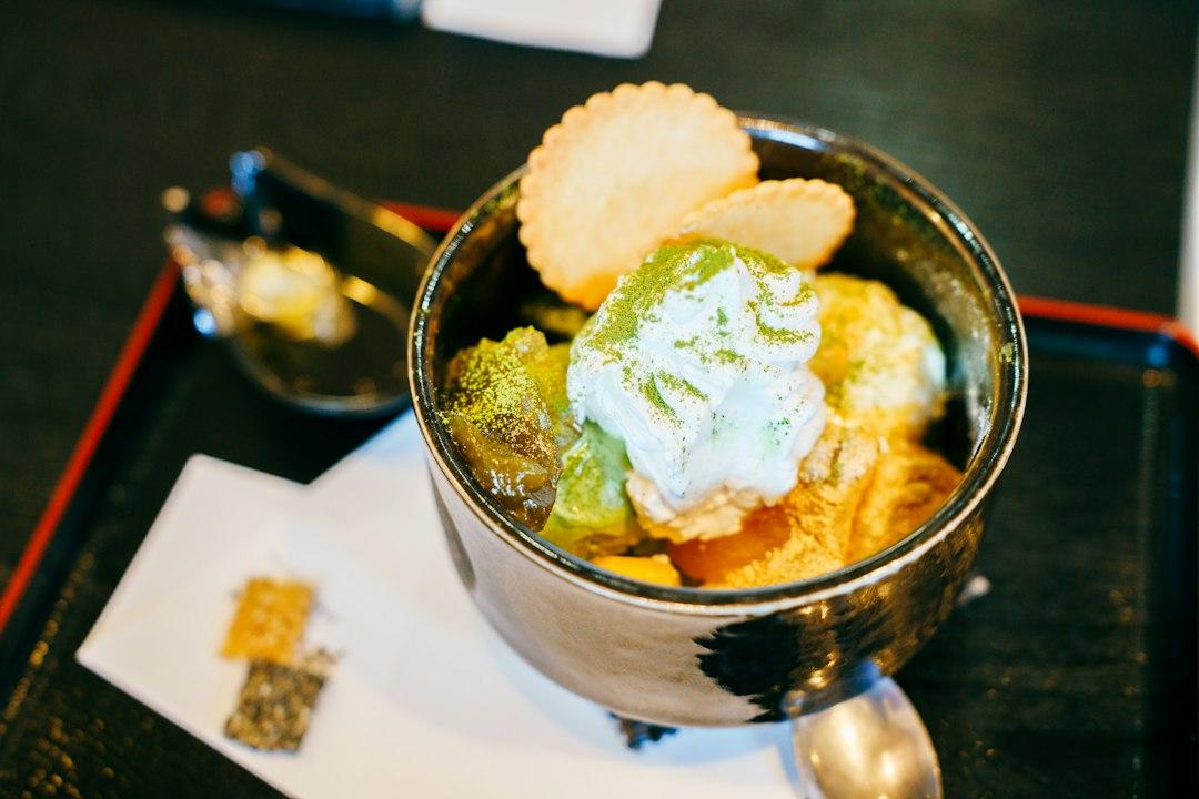 Japanese matcha parfait in a silver bowl topped with whipped cream, matcha powder, and a shell-shaped wafer cookie