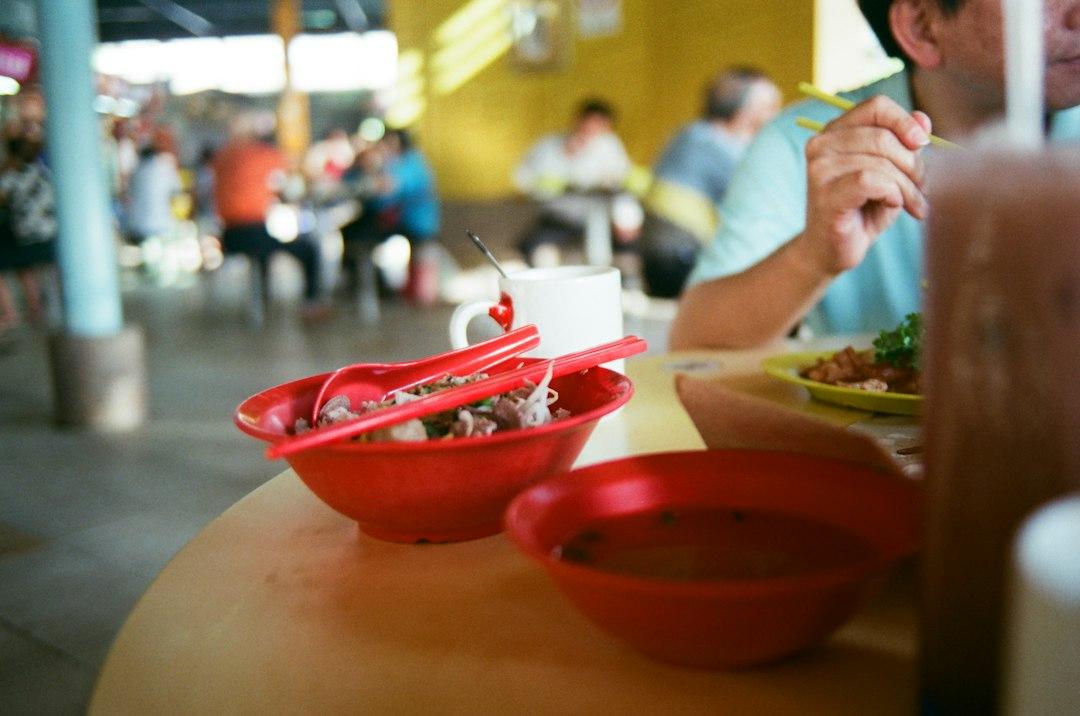 Red bowls of noodle soup on a round table at a busy Singaporean hawker centre, man eating with chopsticks nearby