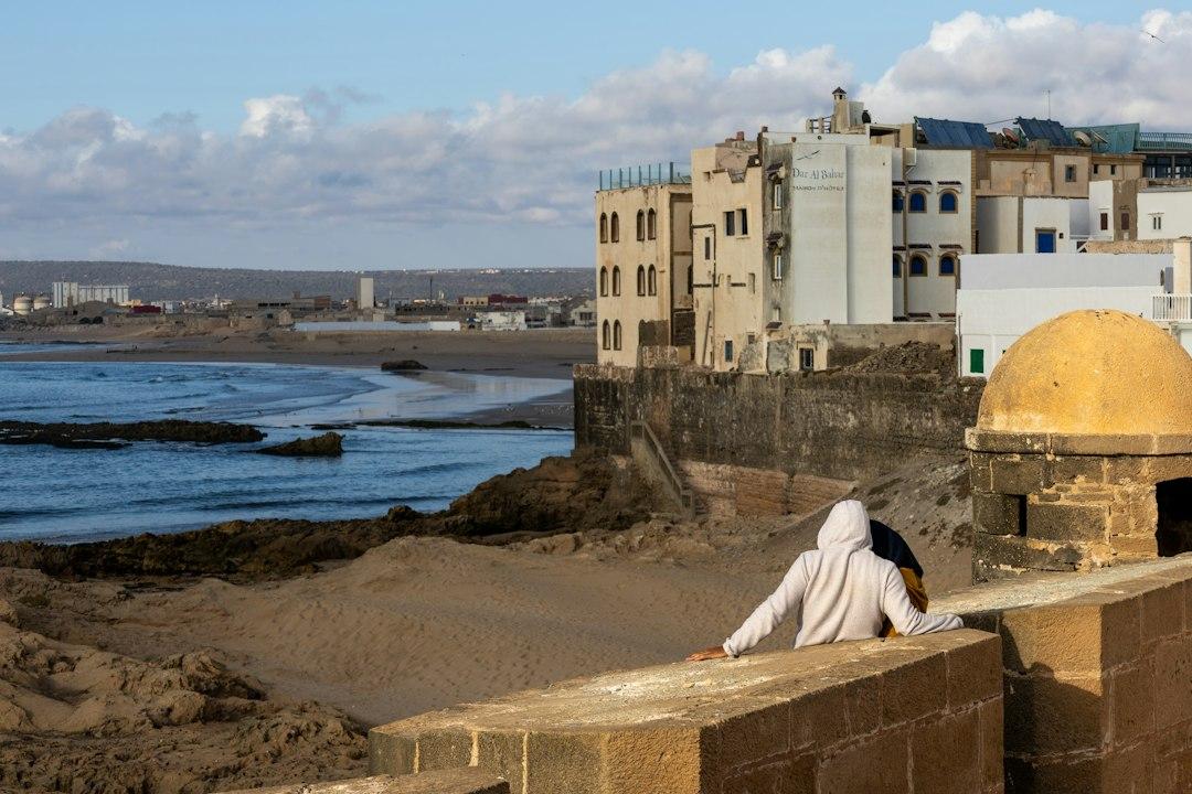 Person in hoodie sitting on Essaouira's ancient stone ramparts overlooking the Atlantic beach and medina buildings