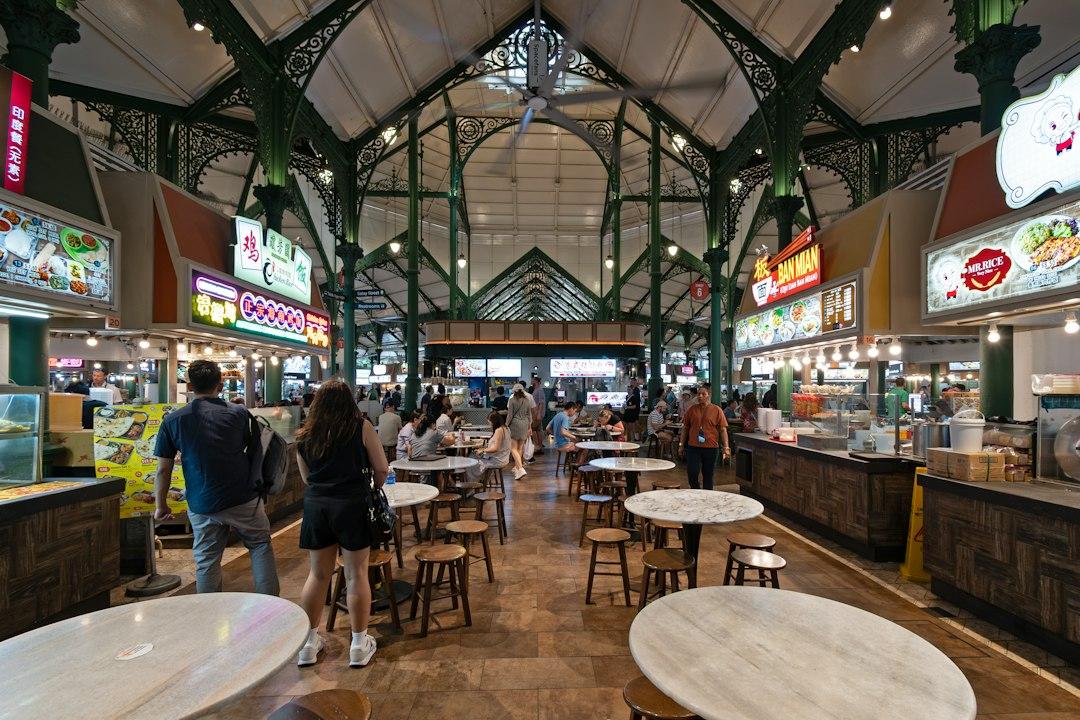 Busy Singapore hawker centre with ornate green iron roof, food stalls, neon signs, and diners at marble tables