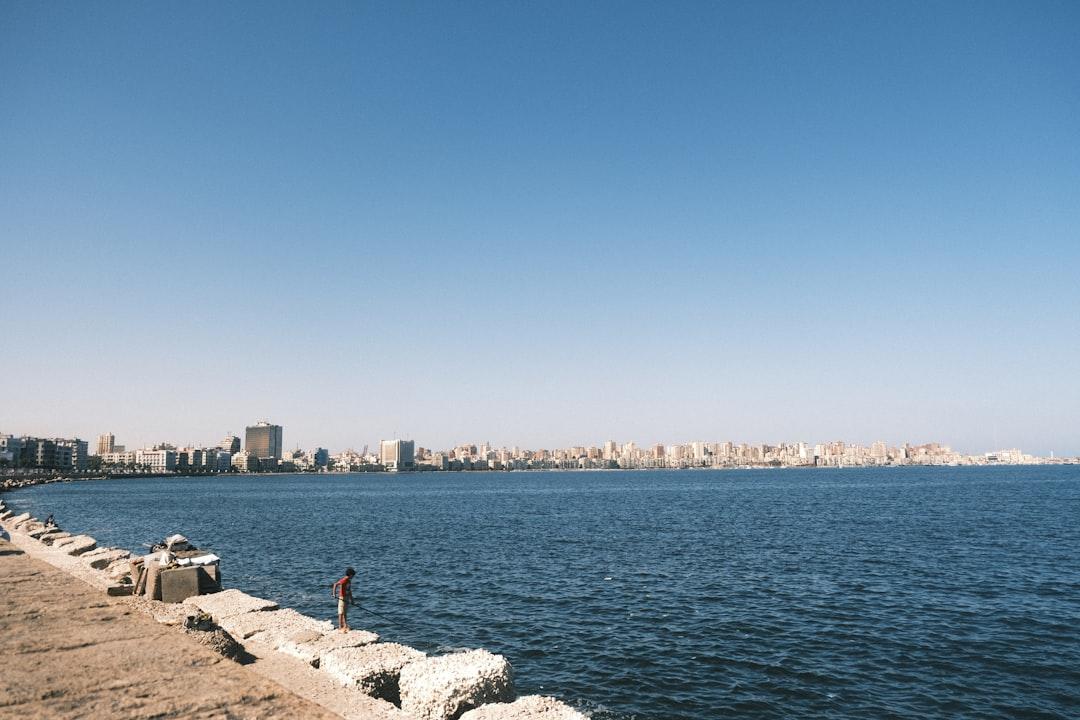 Child fishing on Alexandria's rocky waterfront with the city skyline stretching across the Mediterranean under a clear blue sky