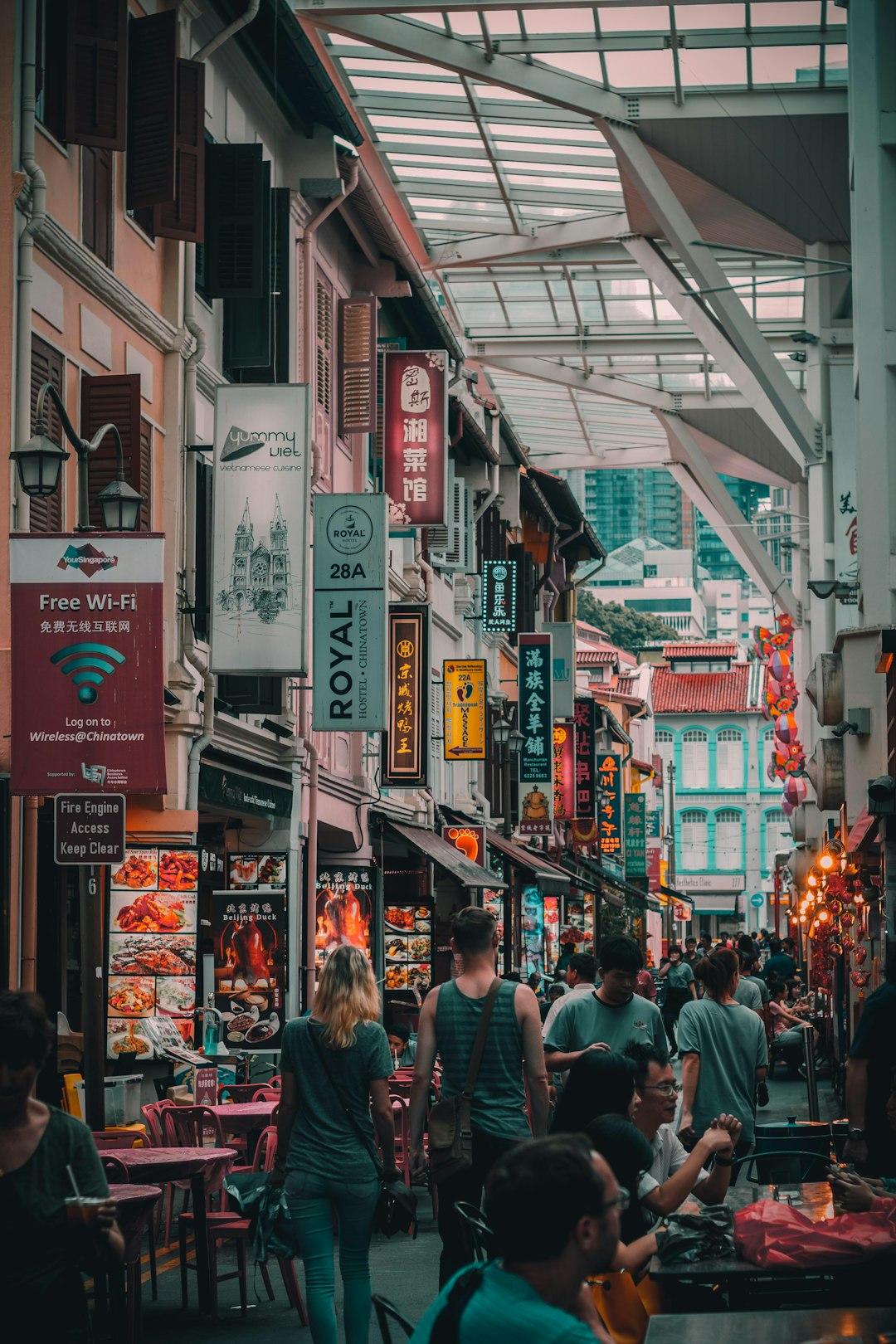 Busy Singapore Chinatown street lined with colorful shophouses, neon signs, and outdoor restaurant tables under a glass canopy