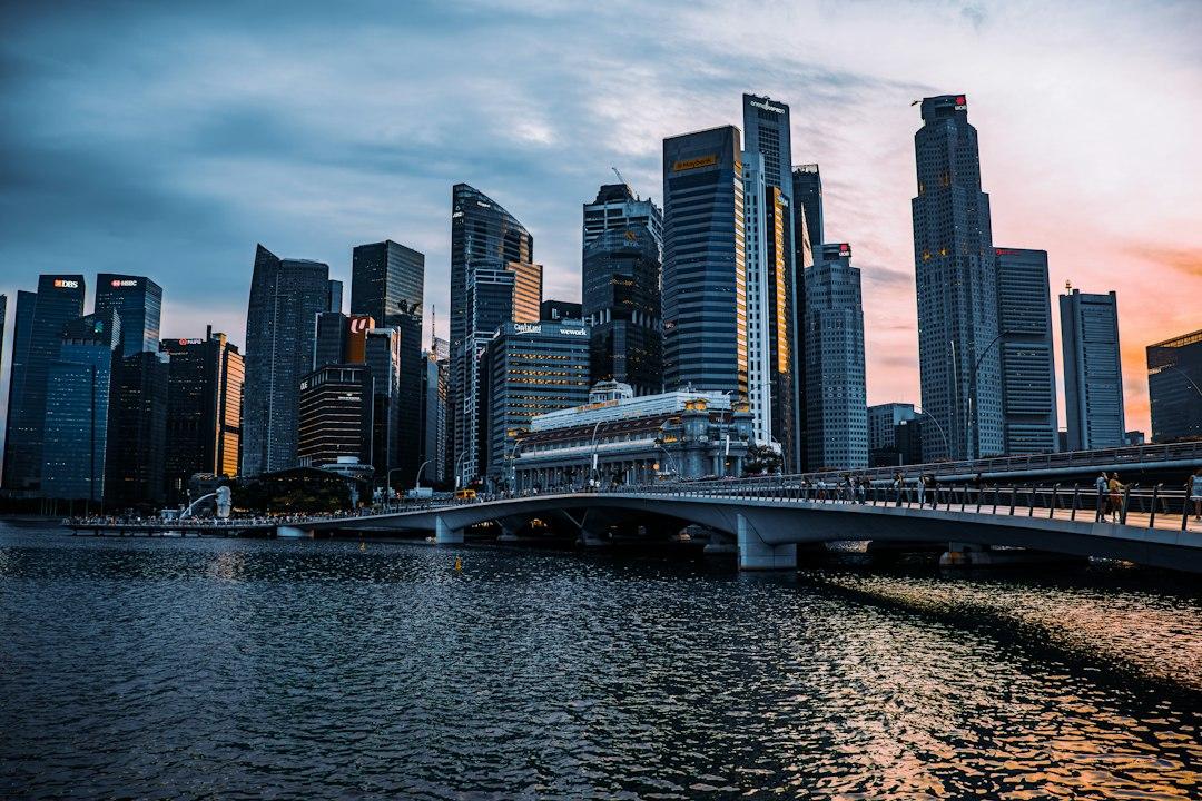 Singapore CBD skyline at dusk with glowing skyscrapers reflected in Marina Bay and a pedestrian bridge in the foreground