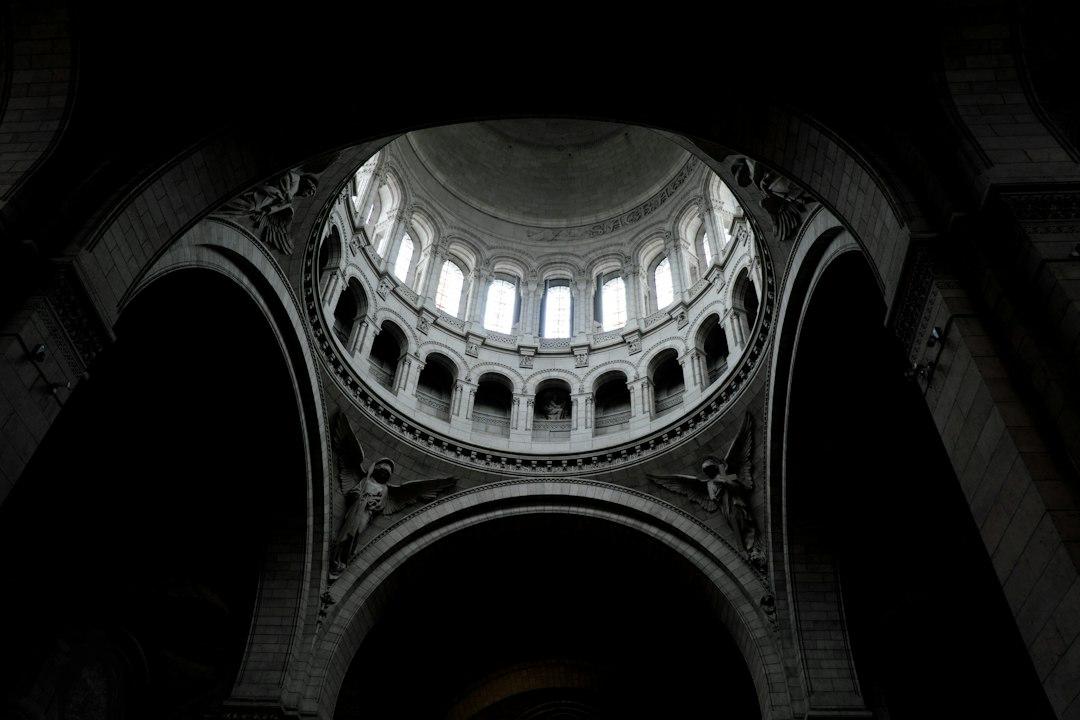 Looking up at the ornate stone dome interior of Sacré-Cœur Basilica, with arched windows and sculpted angels in shadow
