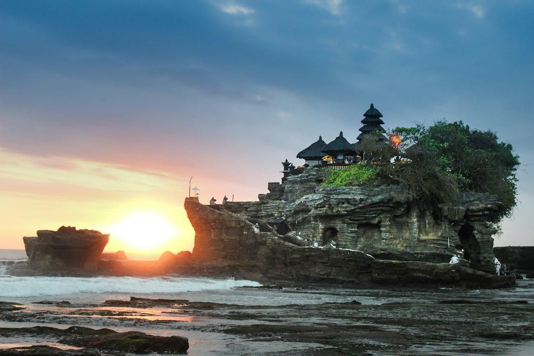 Tanah Lot sea temple perched on a rocky outcrop at sunset, Bali, with tiered pagodas and green trees above crashing waves