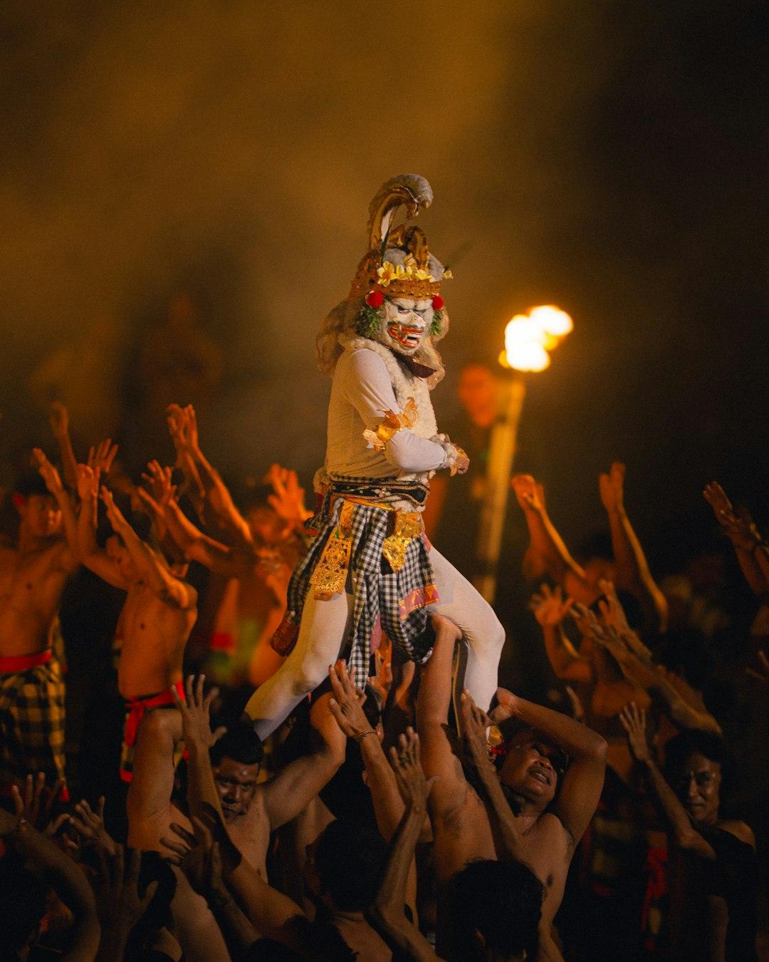 Masked Kecak dancer lifted above crowd of shirtless performers with raised arms during Uluwatu fire dance, Bali