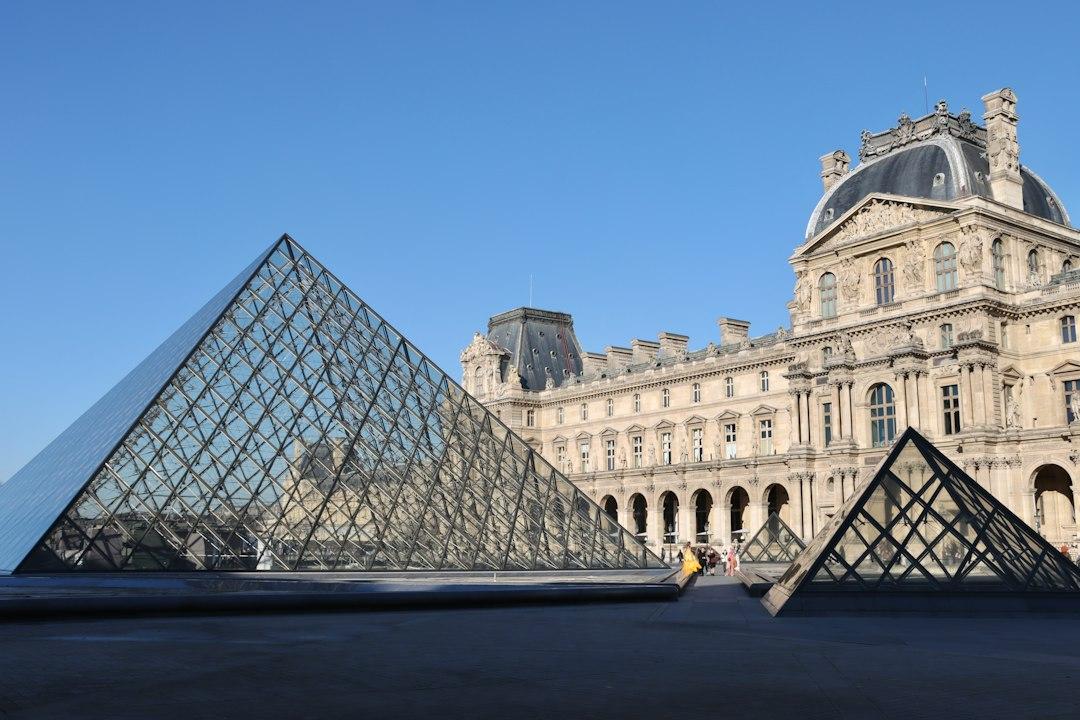 Glass pyramid entrance of the Louvre Museum with the ornate Renaissance palace facade under a clear blue sky, Paris