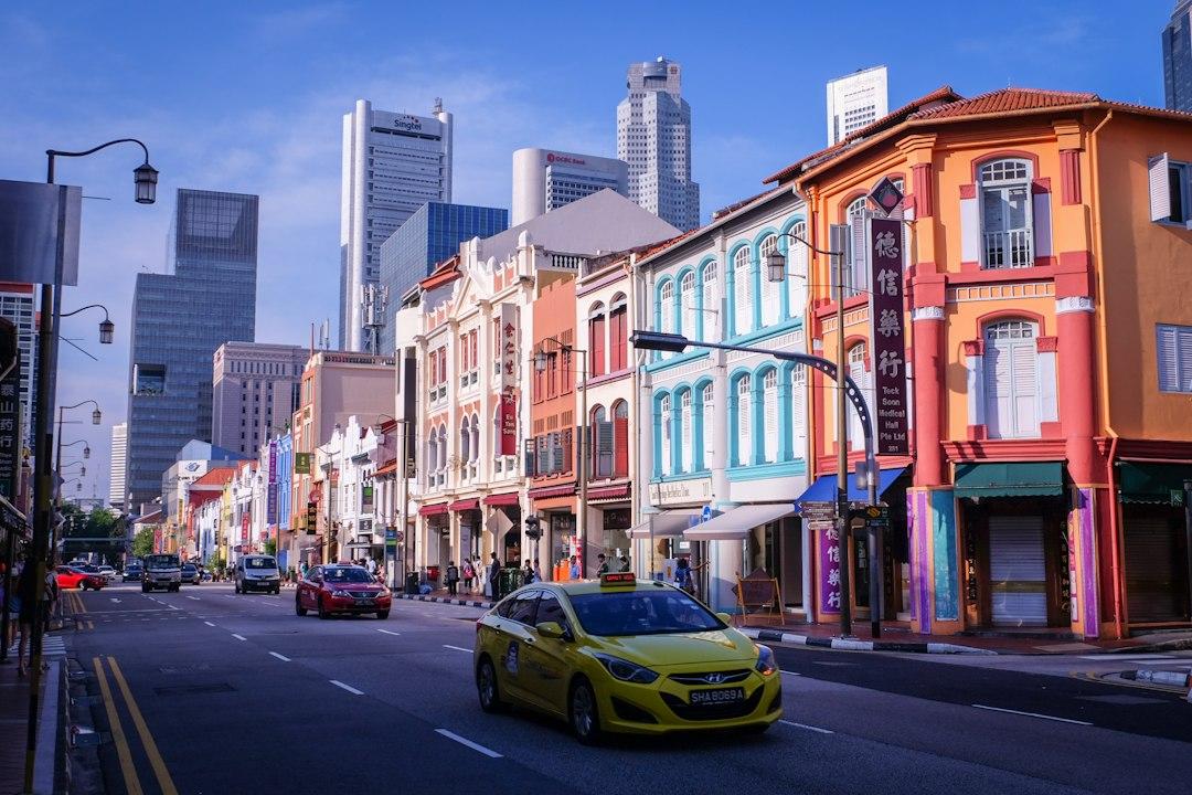 Colorful colonial shophouses lining a busy Singapore Chinatown street with a yellow taxi and modern skyscrapers in the background