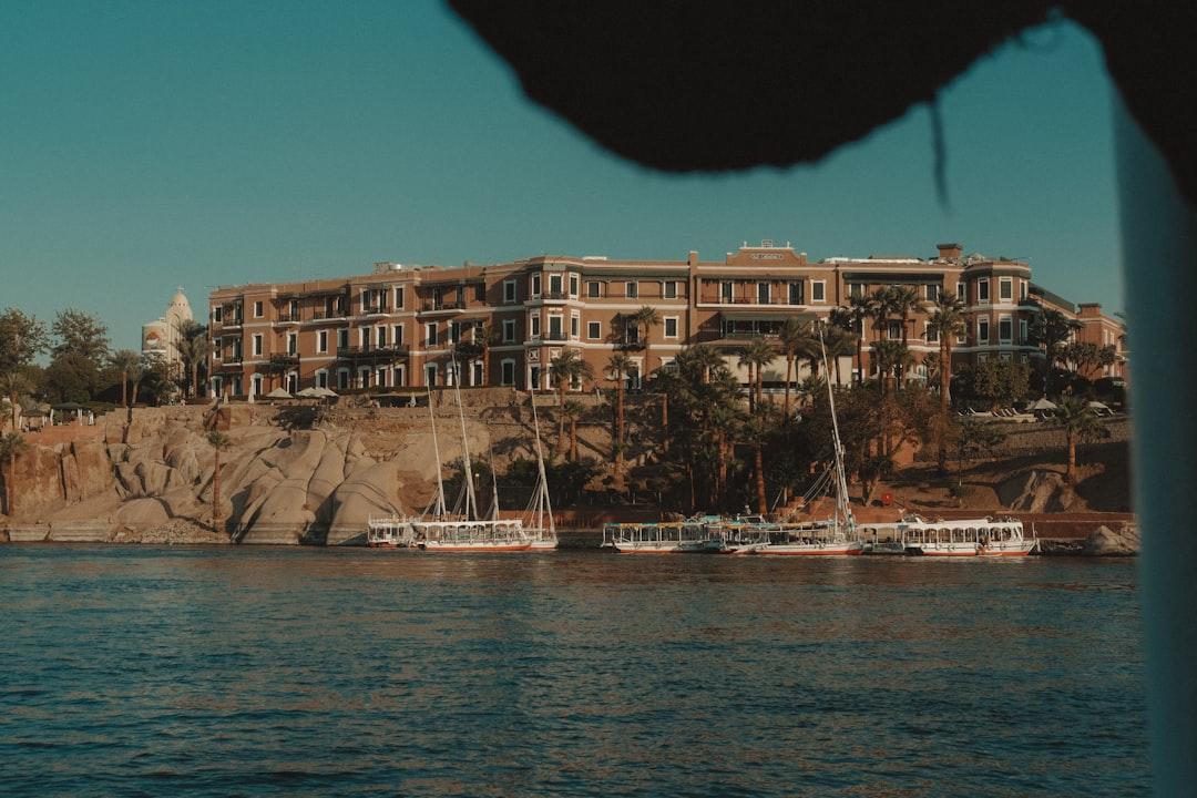 Feluccas and tour boats moored along the Nile beside a grand terracotta hotel on rocky Aswan riverbank