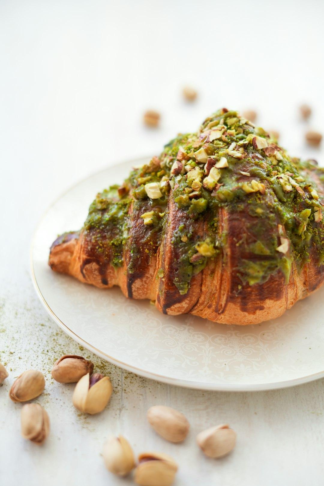 Golden flaky croissant topped with pistachio cream and chopped pistachios on a white plate, surrounded by scattered pistachios