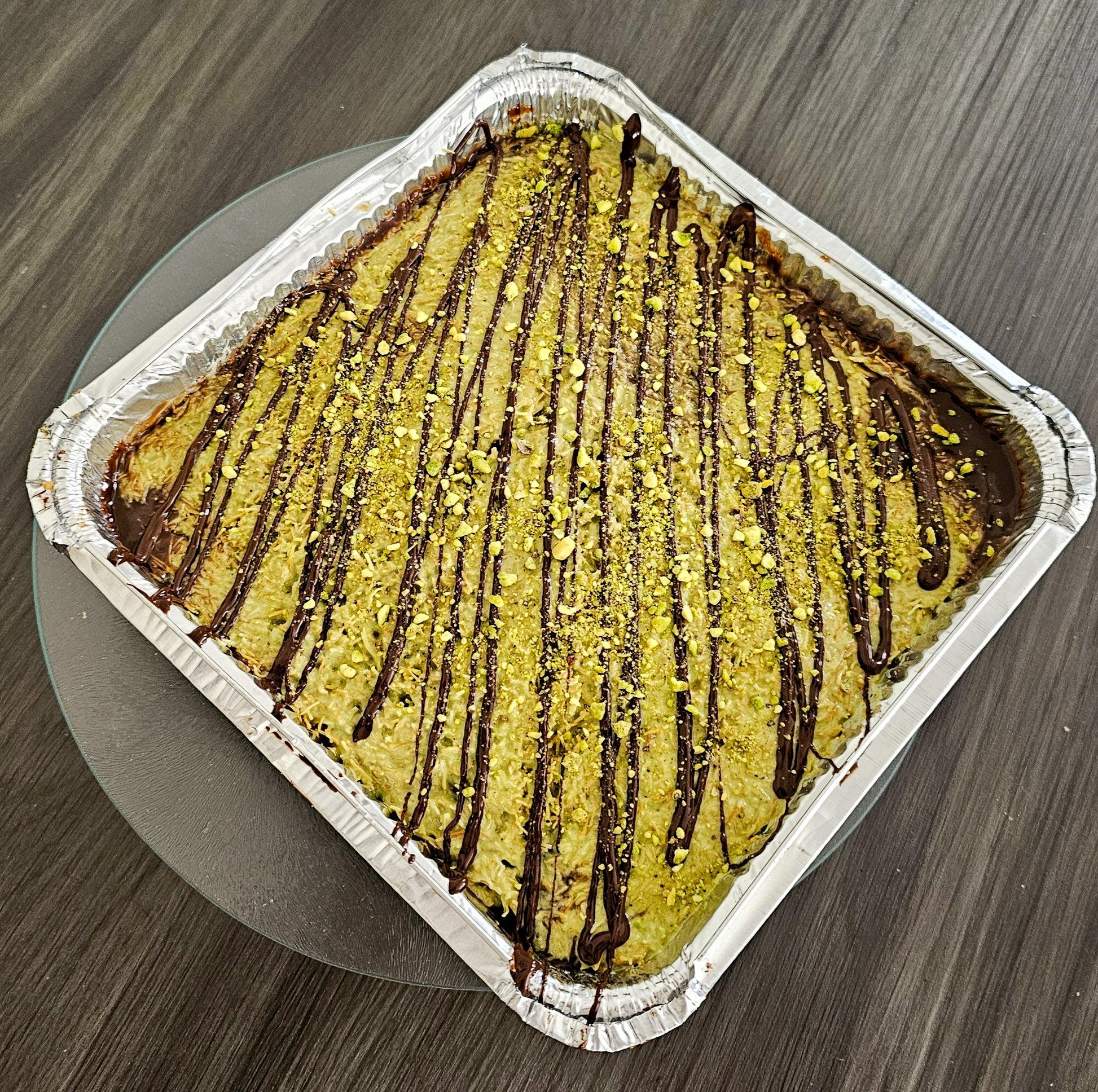 Slice of pistachio kunafa topped with cream and crushed green pistachios on a white plate, with scattered pistachios in background