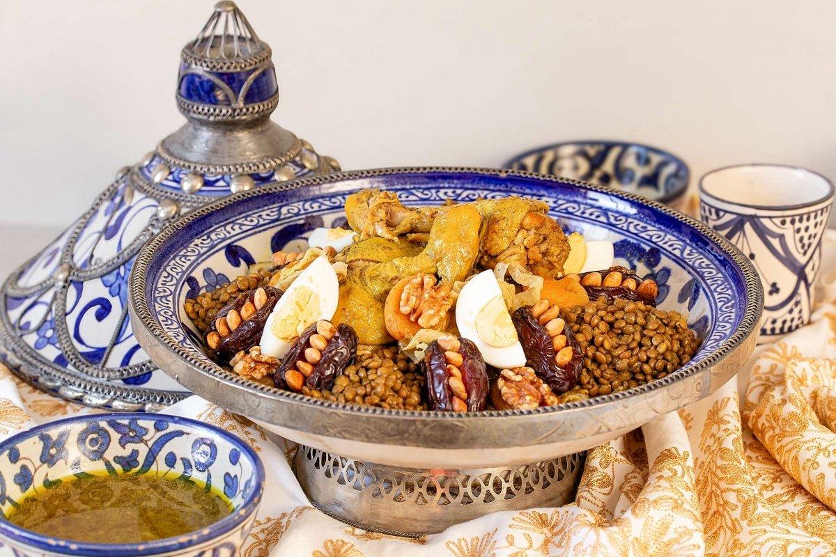 Hands sharing a Moroccan clay tagine dish topped with prunes, nuts, and sesame seeds on a green table