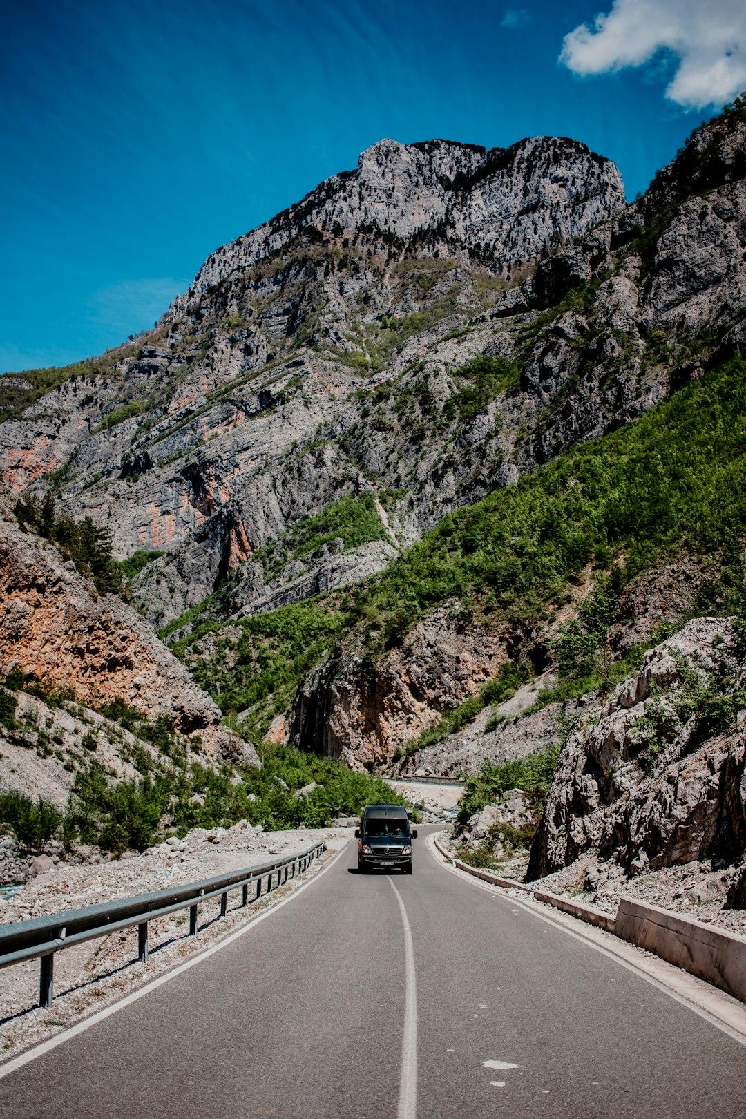 A dark van drives along a narrow mountain road flanked by rocky cliffs and green shrubs under a blue sky in Albania