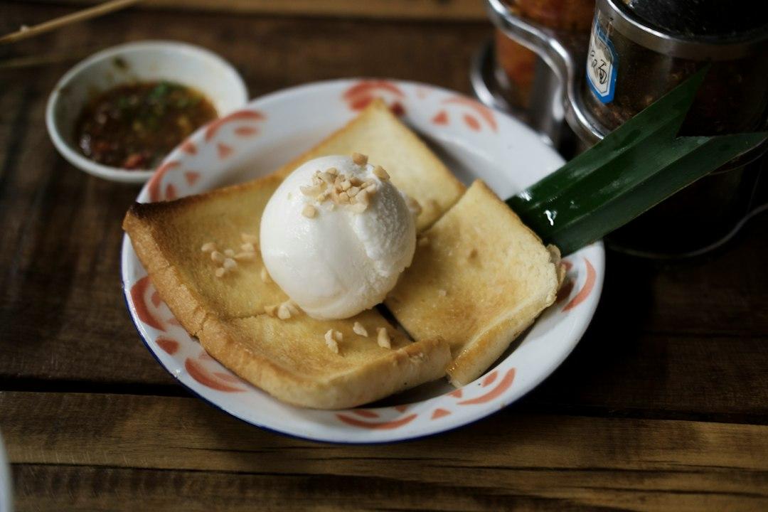 Golden toasted milk bread topped with a scoop of white ice cream and crushed peanuts on a floral plate, wooden table
