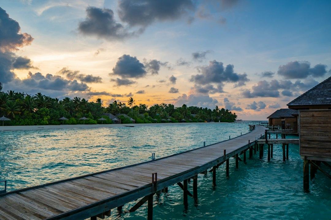 Wooden overwater walkway leading to a Maldives resort villa at sunset, with turquoise lagoon and palm-lined island shore