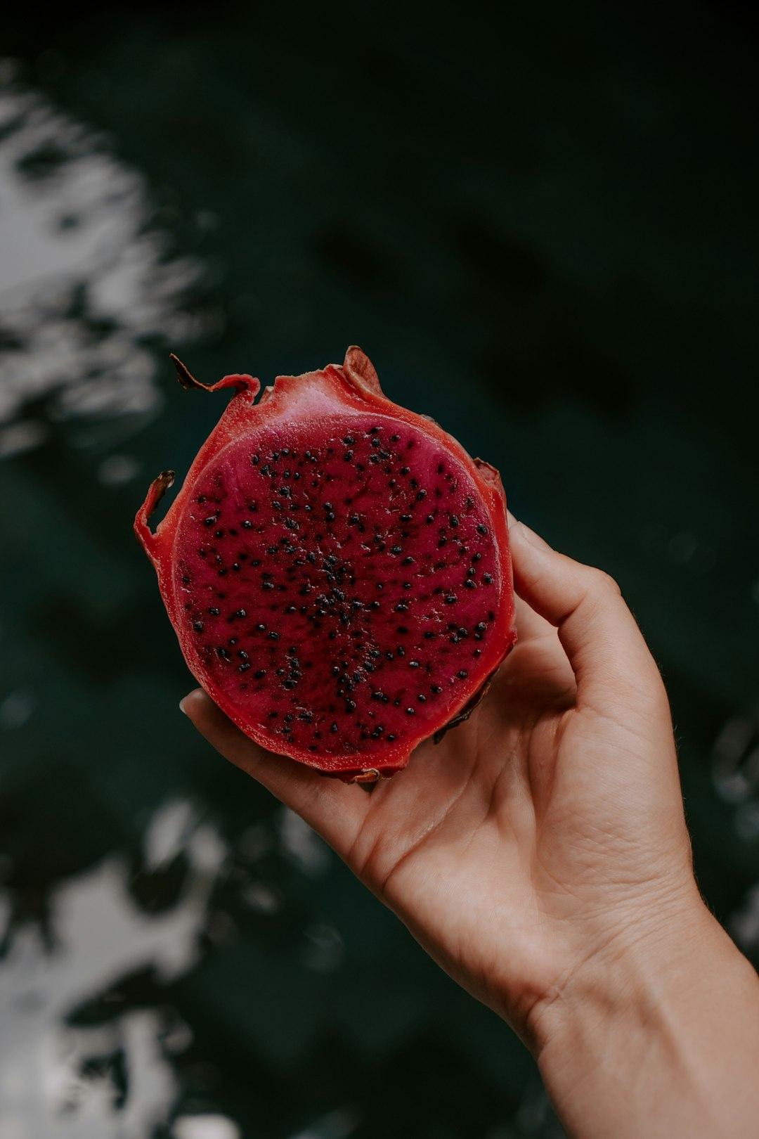 Hand holding a halved red-flesh dragon fruit with vibrant magenta interior and black seeds against dark foliage