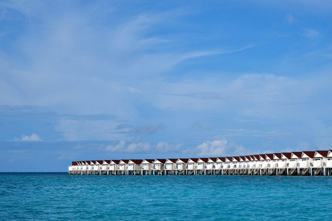 Row of white overwater bungalows with dark roofs on stilts above turquoise Maldives lagoon under blue sky