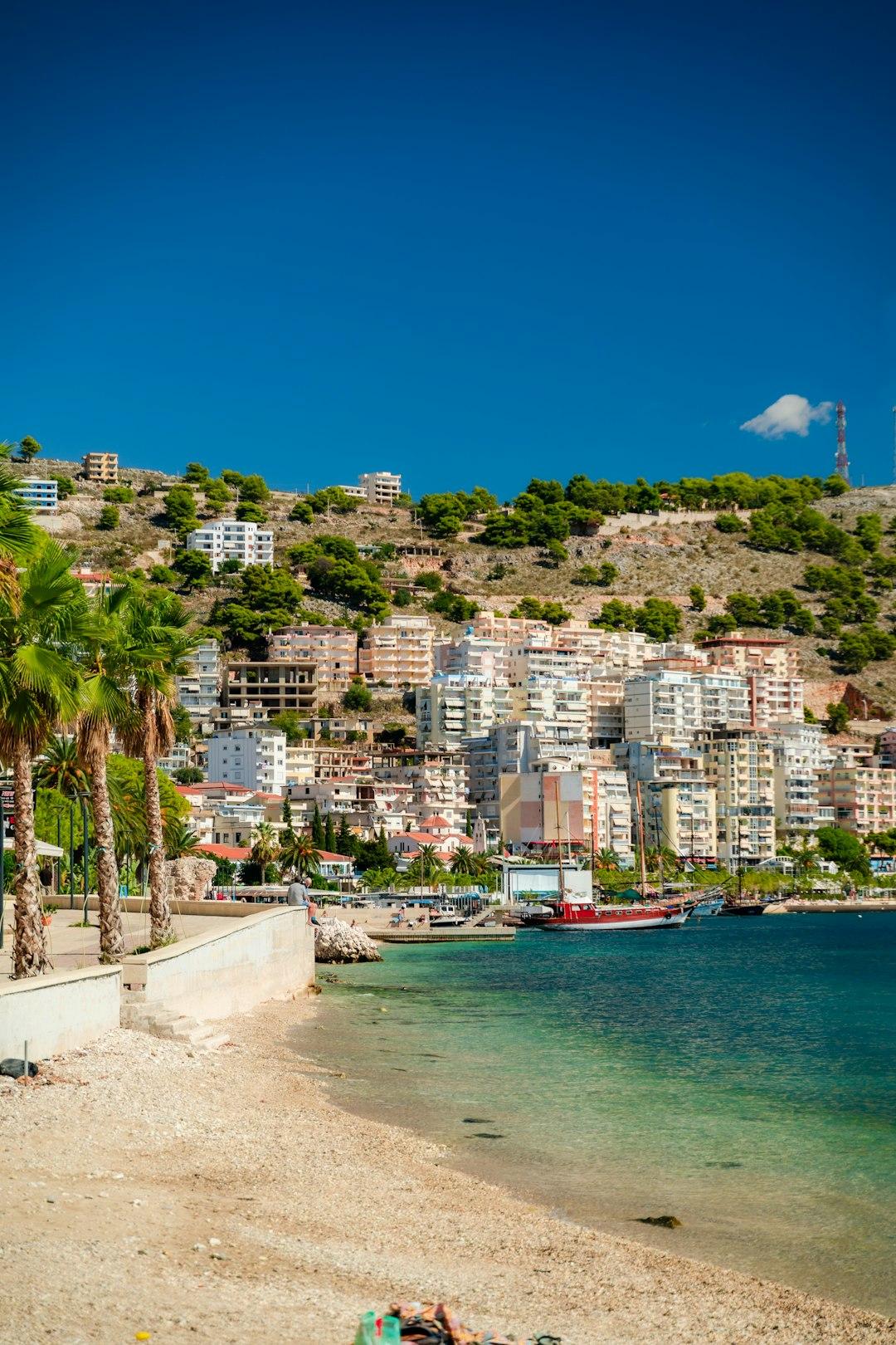 Sandy beach and turquoise sea in Saranda, Albania, with white apartment buildings climbing a pine-covered hillside