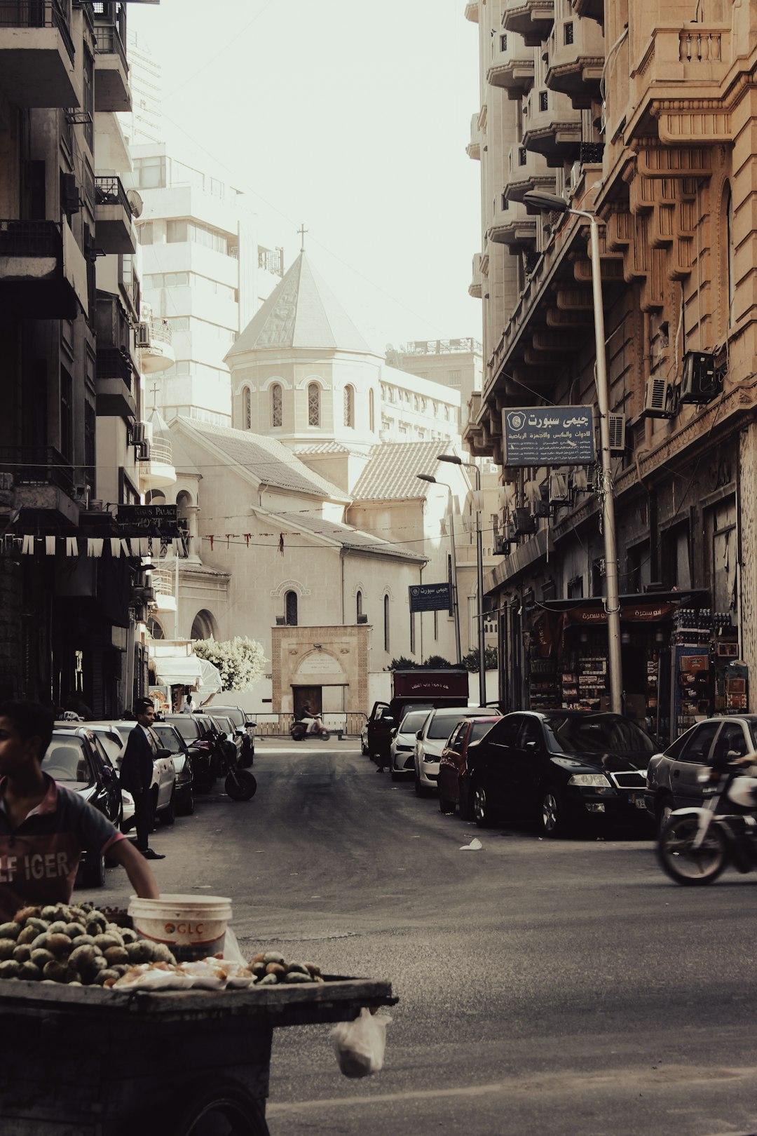 Busy Cairo street lined with parked cars and Arabic shop signs, leading to a stone church with a cross-topped spire