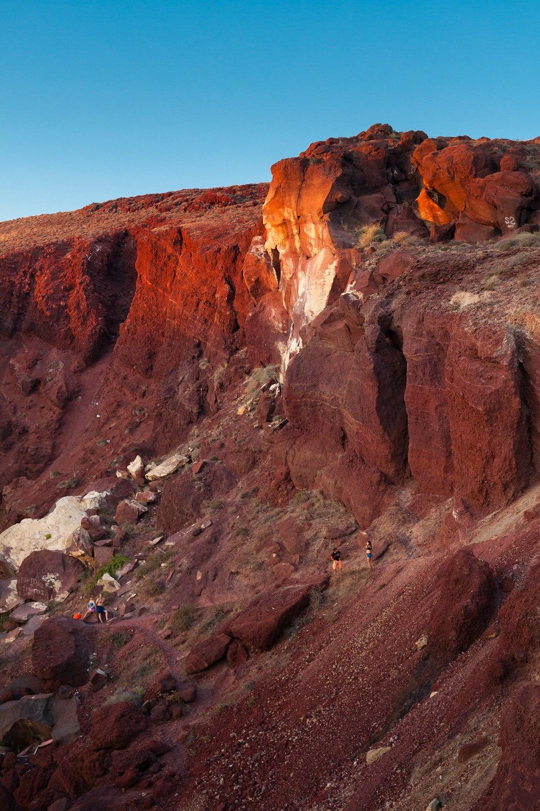 Hikers exploring dramatic red volcanic cliffs with white mineral streaks under a clear blue sky in Santorini