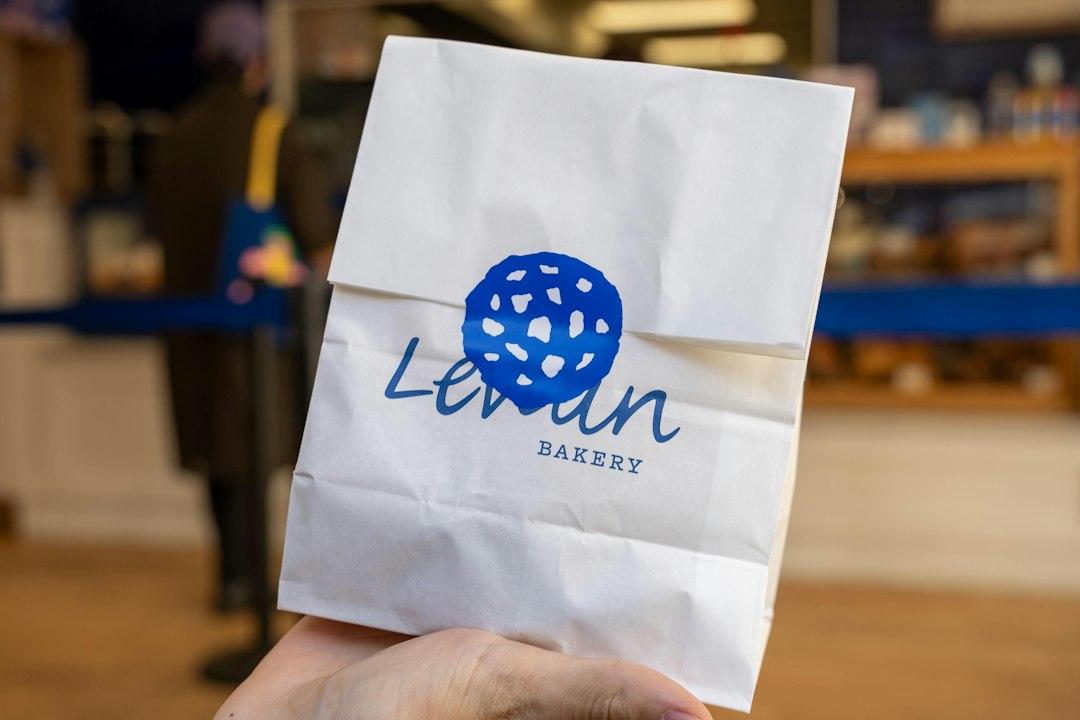 Hand holding a white paper bag printed with the Levain Bakery logo in blue, inside a bakery shop