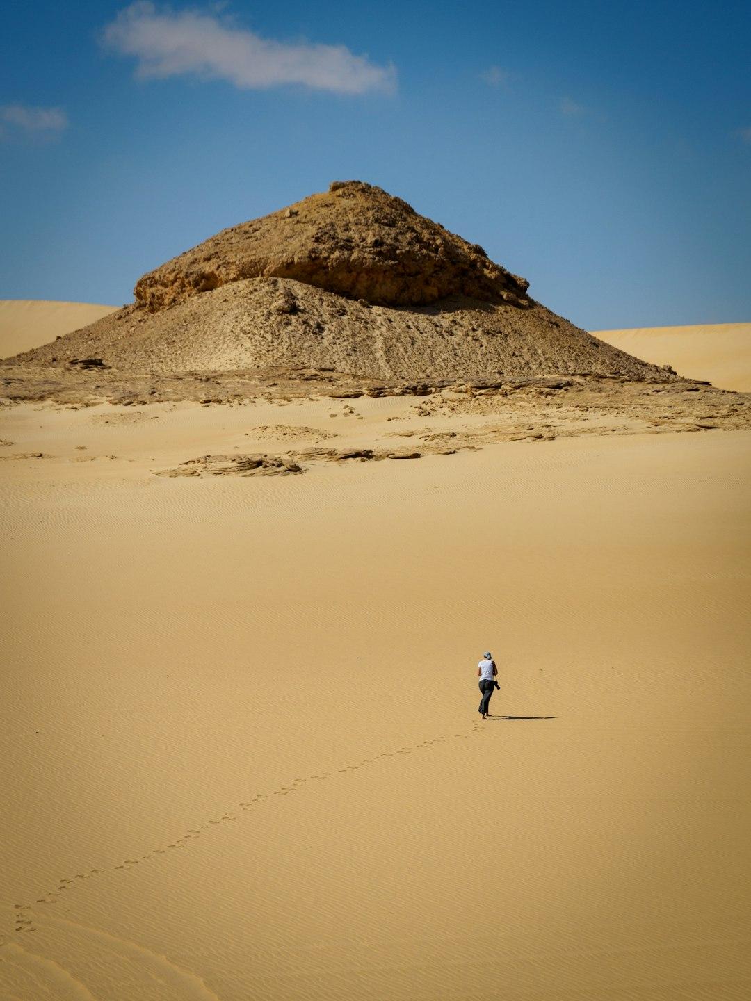 Solo traveler walking across golden sand dunes toward a rocky desert butte under a blue sky in Egypt's Great Sand Sea
