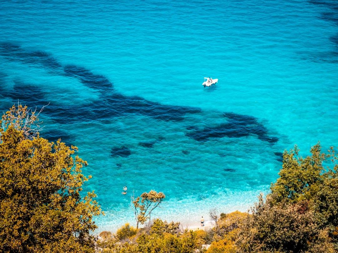 Aerial view of turquoise Ionian Sea cove with a white motorboat, rocky seabed shadows, sandy shore, and green trees in Ksamil, Albania