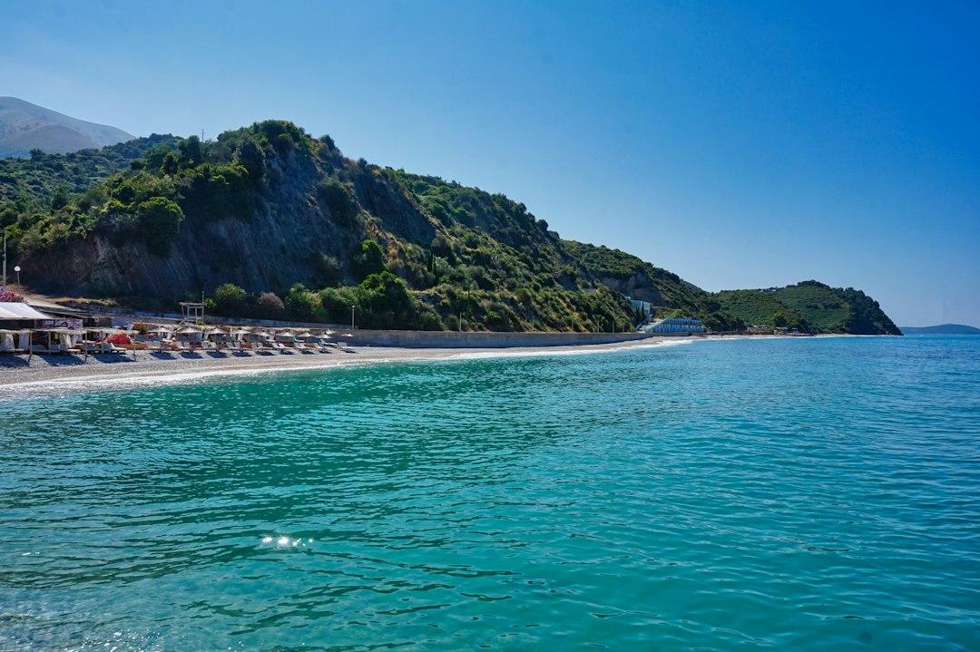 Turquoise waters of an Albanian beach with sun loungers, green hillside, and clear blue sky near Saranda