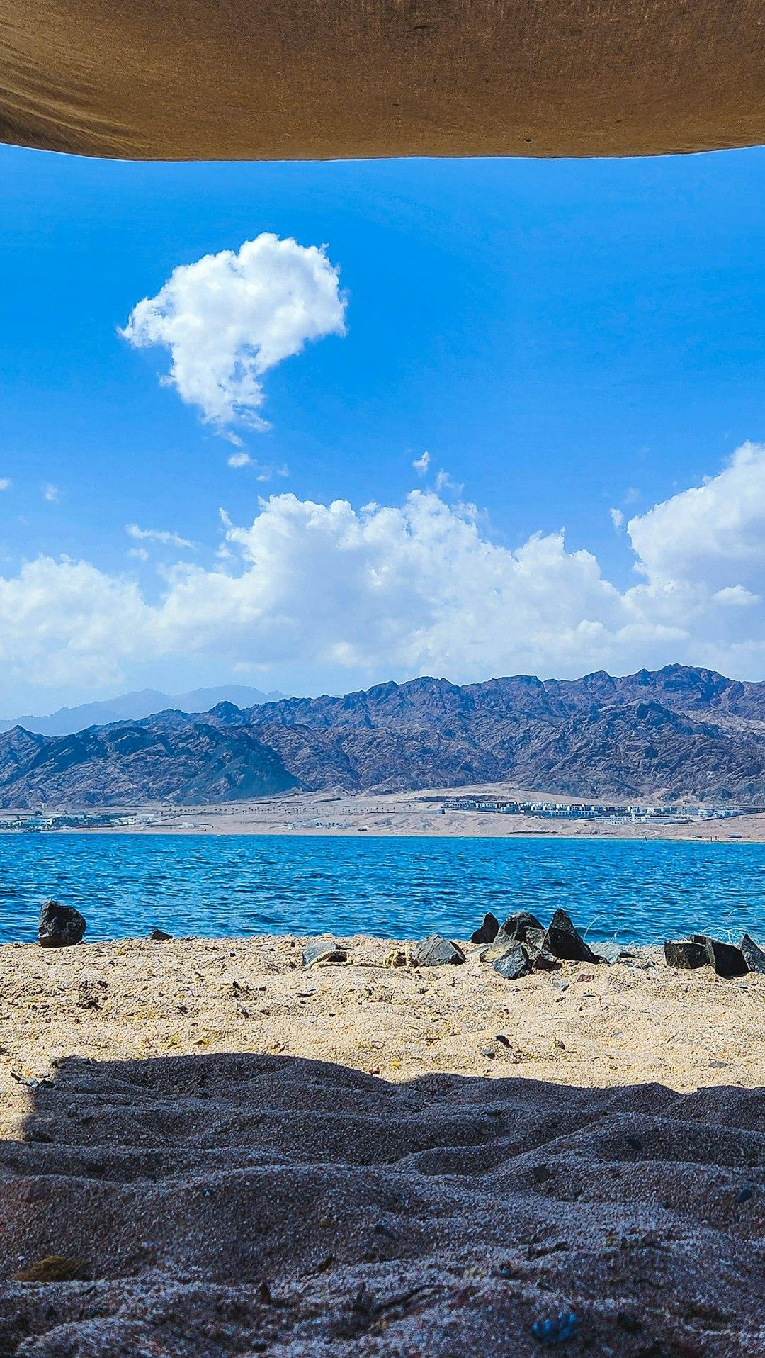Sandy beach with dark rocks framing a blue lagoon, arid mountains, and scattered buildings under a bright sky in El Gouna