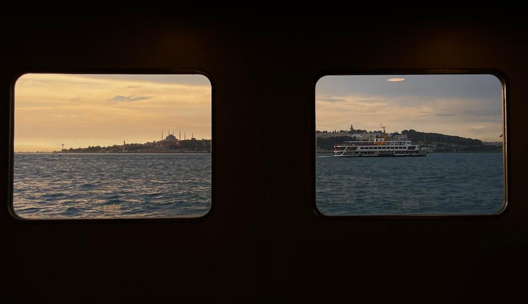 View from ferry portholes across the Bosphorus at sunset, with Istanbul's mosque skyline and a passenger ferry sailing past a green hillside