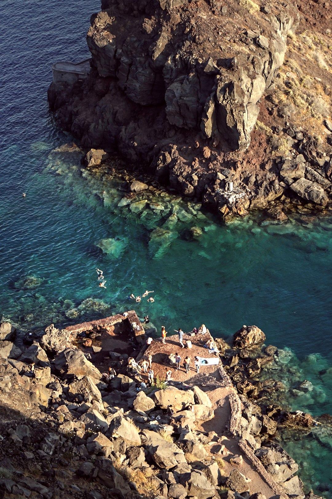 Aerial view of Amoudi Bay, Santorini, with swimmers in turquoise water and visitors resting on volcanic rocks below steep cliffs