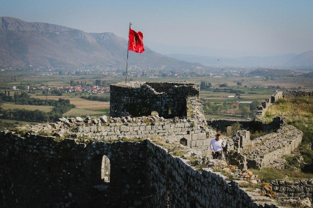 Man walking along ancient stone castle walls with Albanian flag flying, overlooking a green valley and mountains