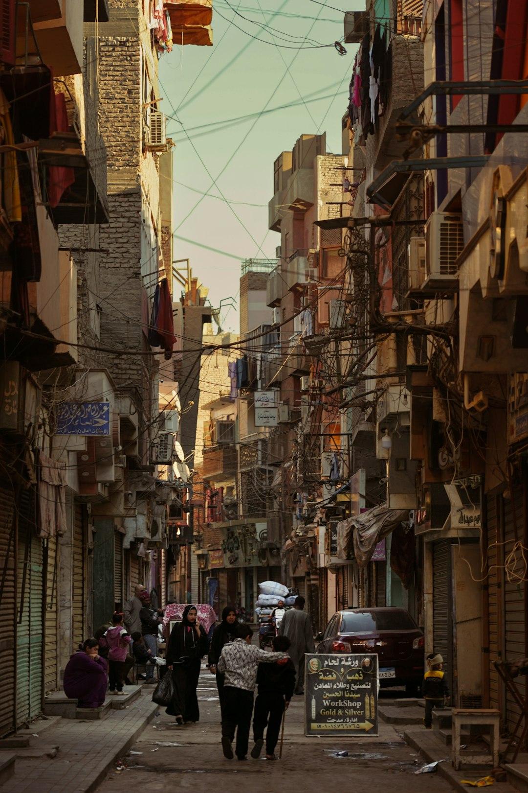 Narrow Cairo residential alley with tangled overhead wires, pedestrians, Arabic shop signs, and laundry hanging from balconies