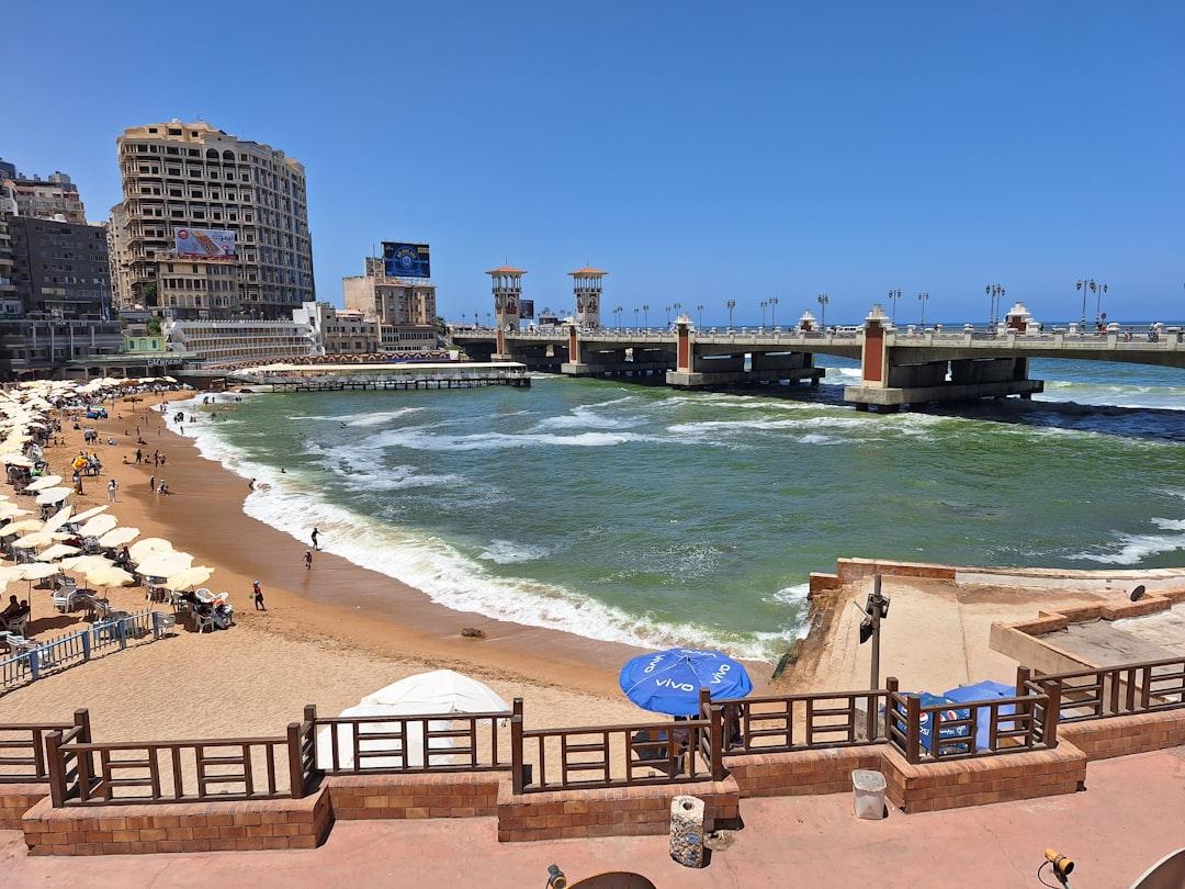 Stanley Beach in Alexandria with turquoise waves, beachgoers, sun umbrellas, and the iconic Stanley Bridge under a clear blue sky