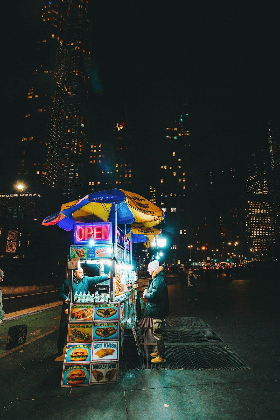 Halal street food cart glowing at night near Brooklyn Bridge, NYC, with a vendor serving a customer under a Sabrett umbrella