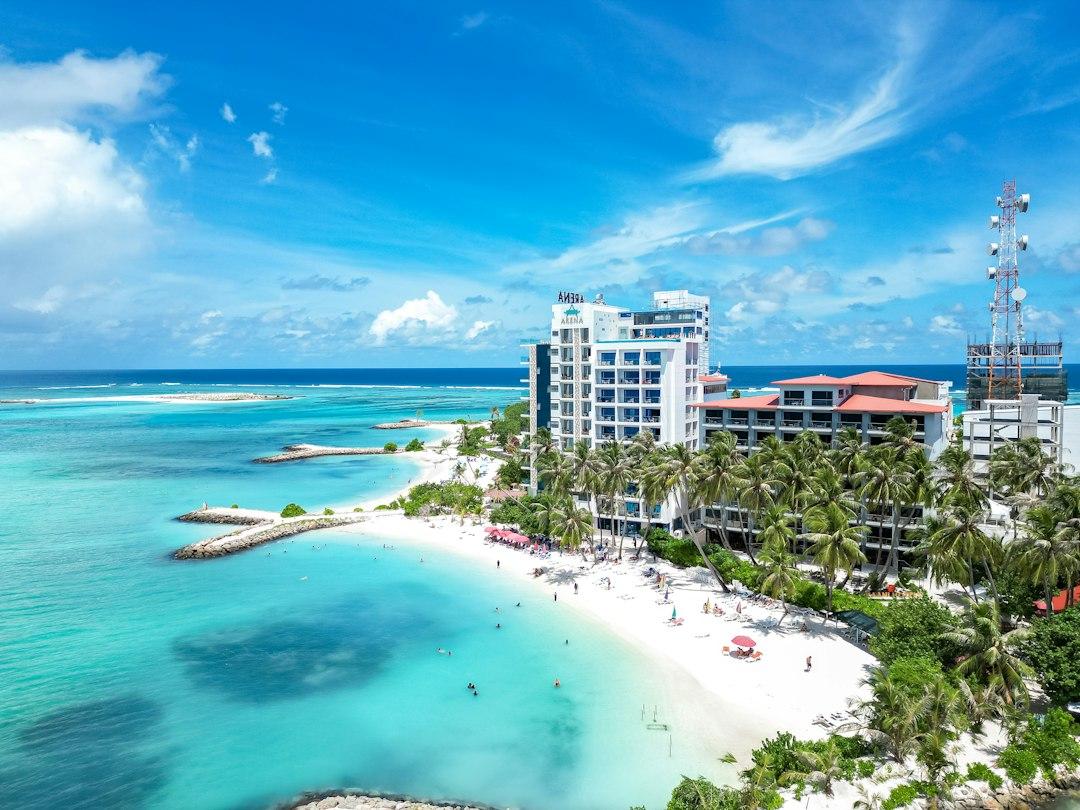 Aerial view of Maafushi island beach with turquoise lagoon, white sand, palm trees, and Arena hotel building, Maldives