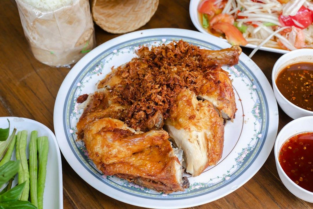 Golden Thai fried chicken topped with crispy garlic on a floral plate, served with sticky rice, papaya salad, and dipping sauces